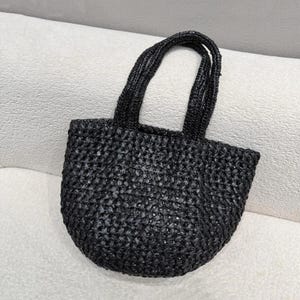 May include: A small, black woven tote bag with two rounded handles. The bag has a rounded bottom and a textured surface. The bag is made of a dark, possibly natural material, and is sitting on a white surface.