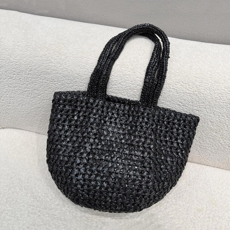 May include: A small, black woven tote bag with two rounded handles. The bag has a rounded bottom and a textured surface. The bag is made of a dark, possibly natural material, and is sitting on a white surface.