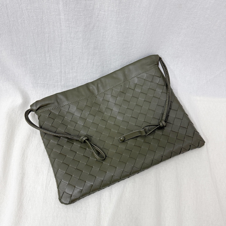 May include: An olive green rectangular clutch bag featuring a woven leather pattern across the entire front surface. The bag includes thin matching leather drawstrings that loop through the top edge and secure with a small, flat leather tab.