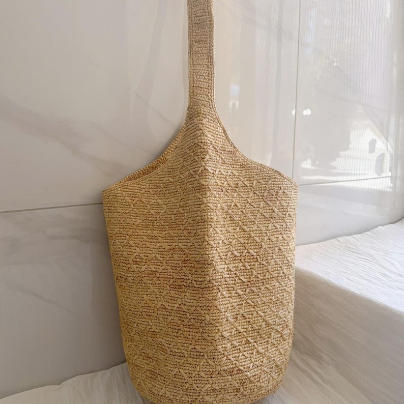 May include: A beige woven tote bag with a long handle. The bag has a diamond pattern and is made of a natural fiber material. The bag is hanging against a white wall.