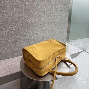 K&ouml;nnte beinhalten: Eine rechteckige senfgelbe Handtasche mit einer weichen, wildlederartigen Textur. Die Tasche verf&uuml;gt &uuml;ber einen Rei&szlig;verschluss an der Oberseite mit zwei Metallschiebern, abgerundete Griffe und strukturierte Kanten. Schlichtes, einfarbiges Design.