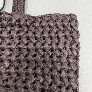 May include: Close-up of a woven tote bag in a muted mauve color. The bag features a tightly woven pattern, with the handle visible at the top. The texture of the material is prominent, suggesting a handmade or artisanal quality.