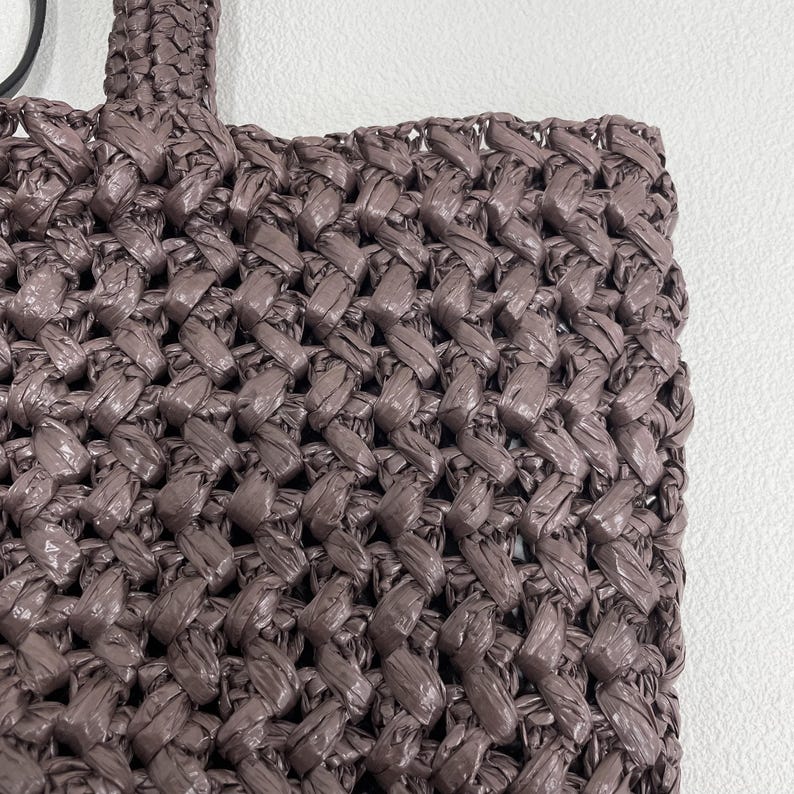 May include: Close-up of a woven tote bag in a muted mauve color. The bag features a tightly woven pattern, with the handle visible at the top. The texture of the material is prominent, suggesting a handmade or artisanal quality.
