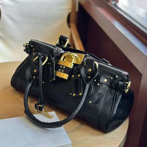 以下が含まれることがあります： 黒い革のハンドバッグ。ゴールドトーンの金具、南京錠の留め具、装飾的なスタッドが特徴です。バッグにはトップハンドルとキーチャームが付いています。バッグは木製の表面に置かれています。