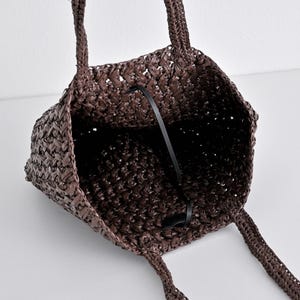 May include: A woven brown tote bag with two handles. The bag has a rectangular shape and a black strap inside. The weave pattern is visible, and the bag appears to be made of a natural material.