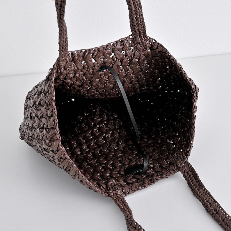 May include: A woven brown tote bag with two handles. The bag has a rectangular shape and a black strap inside. The weave pattern is visible, and the bag appears to be made of a natural material.