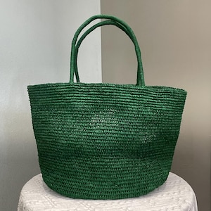 May include: A deep forest green tote bag constructed from woven natural fibers. The bag features a wide, open-top design with a rounded base and two matching green woven handles attached to the top rim, providing a sturdy and consistent texture.