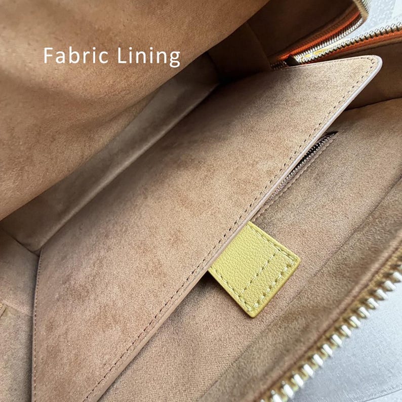 K&ouml;nnte beinhalten: Das Innere einer Tasche mit einem beigefarbenen, wildlederartigen Stofffutter und dem sichtbaren Text Fabric Lining. Es enth&auml;lt eine flache Tasche mit gen&auml;htem Rand und eine kleine, rechteckige, hellgelbe Lederlasche an der Taschen&ouml;ffnung.