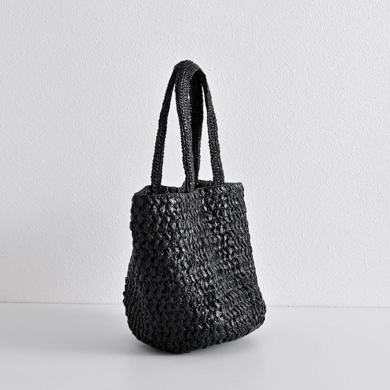 May include: A black woven tote bag with two handles. The bag has a textured appearance and a rounded bottom. The handles are also woven and appear sturdy. The bag is sitting on a white surface.