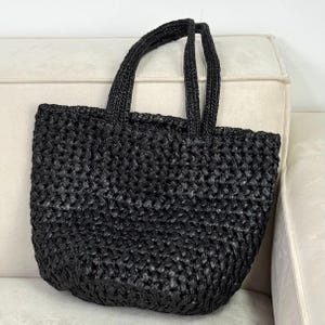May include: A black woven tote bag with two handles. The bag has a rectangular shape and a textured surface. The handles are also black and woven. The bag is sitting on a light-colored surface.