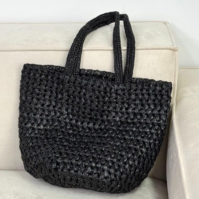 May include: A black woven tote bag with two handles. The bag has a rectangular shape and a textured surface. The handles are also black and woven. The bag is sitting on a light-colored surface.