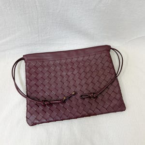 May include: A deep burgundy rectangular pouch featuring an intricate woven leather pattern across the front surface. The bag includes thin, matching leather drawstring cords that loop along the sides and are secured with small gold-toned metal hardware.