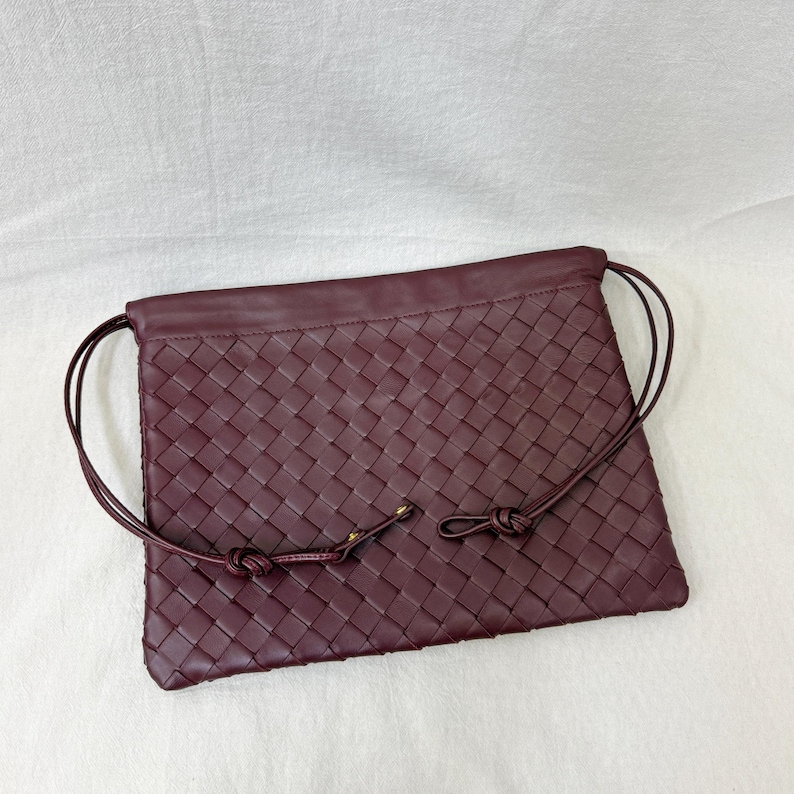 May include: A deep burgundy rectangular pouch featuring an intricate woven leather pattern across the front surface. The bag includes thin, matching leather drawstring cords that loop along the sides and are secured with small gold-toned metal hardware.