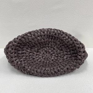 May include: A dark brown, woven raffia hat with a rounded shape. The hat is made of tightly woven strands, creating a textured surface. The hat is a neutral color and could be worn in the summer.