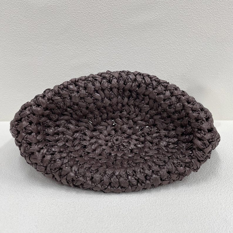 May include: A dark brown, woven raffia hat with a rounded shape. The hat is made of tightly woven strands, creating a textured surface. The hat is a neutral color and could be worn in the summer.