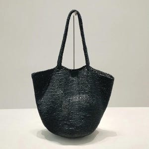 Può includere: Una borsa tote nera intrecciata con due manici. La borsa ha un fondo arrotondato e un'ampia apertura. Il materiale sembra essere rafia a trama fitta o simile. La borsa è su una superficie bianca.