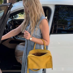 K&ouml;nnte beinhalten: Eine leuchtend gelbe Wildlederhandtasche mit strukturierter rechteckiger Form, einem Tragegriff und einem Rei&szlig;verschluss. Die Tasche verf&uuml;gt &uuml;ber ein glattes Lederdetail an der unteren Ecke und lange, d&uuml;nne Wildlederfransen an den Seiten.