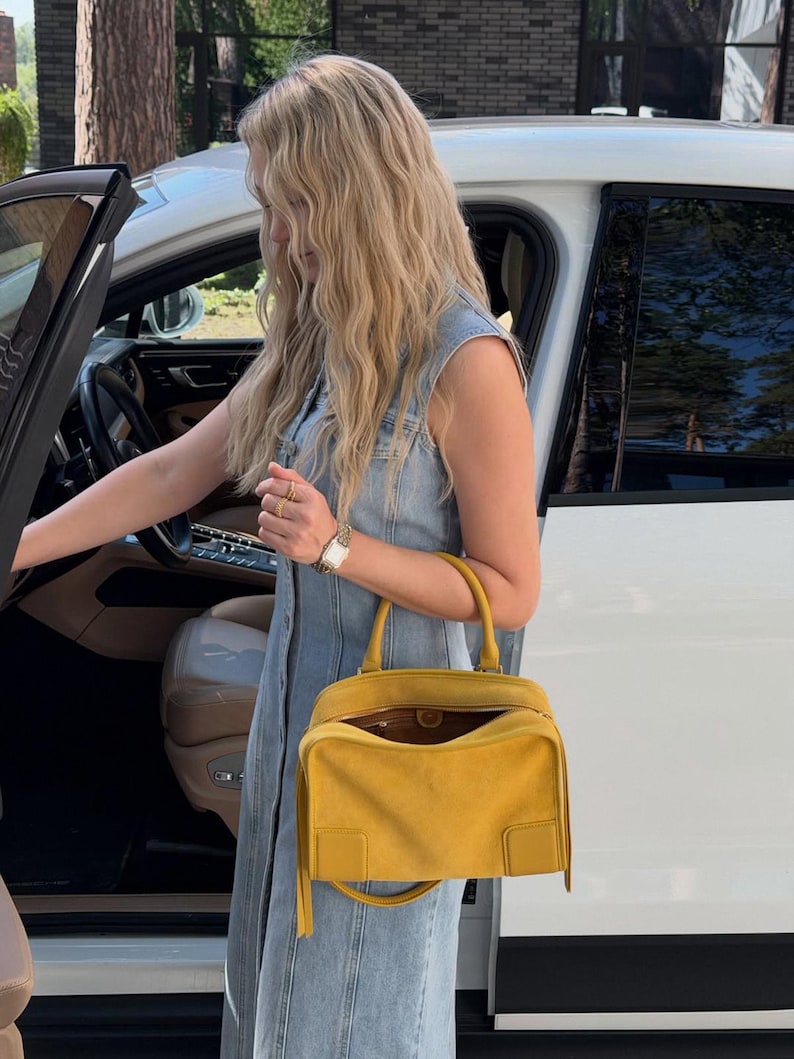 K&ouml;nnte beinhalten: Eine leuchtend gelbe Wildlederhandtasche mit strukturierter rechteckiger Form, einem Tragegriff und einem Rei&szlig;verschluss. Die Tasche verf&uuml;gt &uuml;ber ein glattes Lederdetail an der unteren Ecke und lange, d&uuml;nne Wildlederfransen an den Seiten.