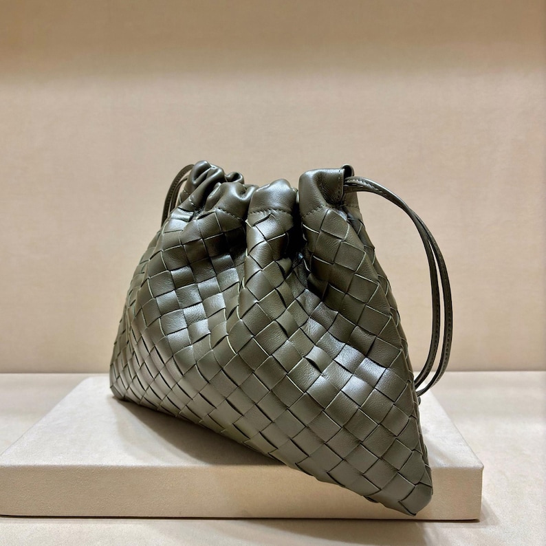 May include: An olive green leather pouch featuring a woven intrecciato pattern. The bag has a gathered top opening with thin leather drawstring cords on each side, creating a soft, rounded silhouette with a smooth, uniform matte finish.