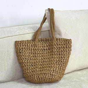 May include: A woven tote bag in a natural tan color. The bag has two handles and a textured, open weave design. The bag is sitting on a white surface.