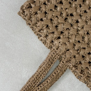 May include: Close-up of a woven tote bag in a light brown color. The bag features a textured, open weave design. The straps are also woven and appear to be sturdy. The bag is likely made of natural fibers, perfect for carrying items.