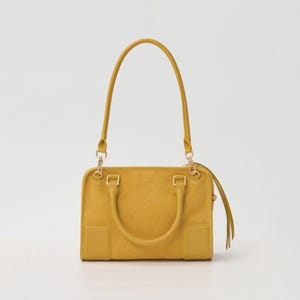 K&ouml;nnte beinhalten: Eine senfgelbe Handtasche mit rechteckigem Korpus, zwei kurzen Tragegriffen und einem langen, abnehmbaren Schulterriemen. Die Tasche verf&uuml;gt &uuml;ber goldfarbene Metallbeschl&auml;ge, darunter Clips und Ringe, sowie einen Rei&szlig;verschluss mit langen Lederzippern.