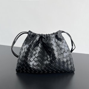 May include: A black handbag featuring a woven leather exterior with a square grid pattern. The bag has a gathered, drawstring-style top closure and two thin, rounded black leather shoulder straps extending from the sides of the main body.