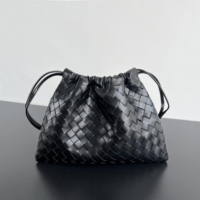 May include: A black handbag featuring a woven leather exterior with a square grid pattern. The bag has a gathered, drawstring-style top closure and two thin, rounded black leather shoulder straps extending from the sides of the main body.