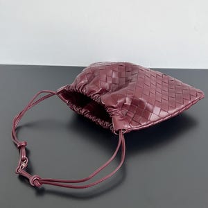 May include: A deep burgundy pouch bag featuring a woven leather exterior with a square grid pattern. The bag has a gathered opening at the top and a thin, matching burgundy leather drawstring strap with a knotted detail for closure and carrying.