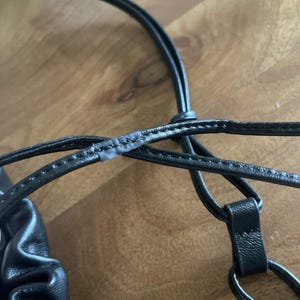 May include: A black bag featuring thin, rounded straps with visible stitching along the edges. The straps are connected by a circular ring attachment, and the bag material shows a gathered, textured surface detail near the strap connection point.