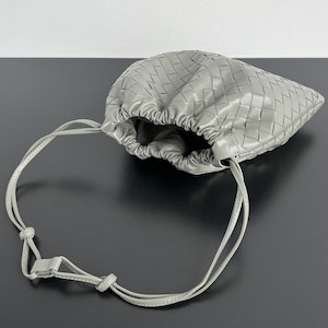 May include: A sage green pouch bag featuring a woven leather exterior with a gathered drawstring opening. The bag includes a long, thin, adjustable shoulder strap with a sliding knot mechanism, creating a soft, rounded shape with a textured finish.