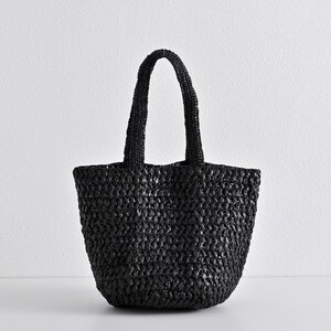 May include: A black woven tote bag with two handles. The bag has a rounded bottom and a textured surface. The handles are thick and appear to be made of the same material as the bag. The bag is sitting on a white surface.