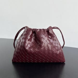 May include: A deep burgundy handbag featuring a woven leather exterior with a grid pattern. The bag has a gathered, drawstring-style top closure and two thin, matching leather shoulder straps extending from the sides of the pouch-shaped body.