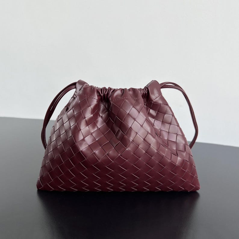 May include: A deep burgundy handbag featuring a woven leather exterior with a grid pattern. The bag has a gathered, drawstring-style top closure and two thin, matching leather shoulder straps extending from the sides of the pouch-shaped body.