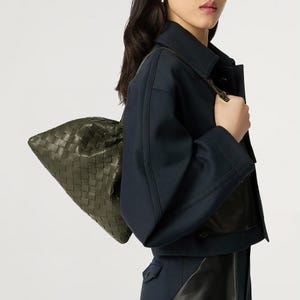 May include: A dark olive green woven leather shoulder bag with a soft, slouchy silhouette and a distinct grid-like pattern. The bag is paired with a dark navy blue jacket featuring wide sleeves and structured seams, worn over a dark garment.
