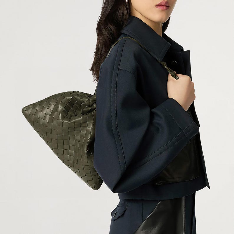 May include: A dark olive green woven leather shoulder bag with a soft, slouchy silhouette and a distinct grid-like pattern. The bag is paired with a dark navy blue jacket featuring wide sleeves and structured seams, worn over a dark garment.