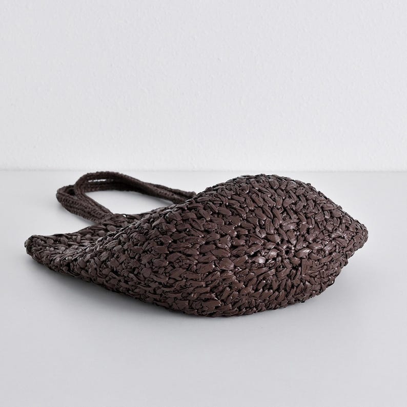 May include: A dark brown woven tote bag with two handles. The bag is made of a tightly woven material, creating a textured surface. The bag is lying on its side, showcasing its shape and construction. The bag is a neutral color and suitable for various uses.