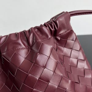 May include: A deep burgundy handbag featuring a woven leather construction with a smooth, matte finish. The bag has a gathered, drawstring-style top closure with a thin leather strap extending from the side, showcasing a detailed grid pattern.