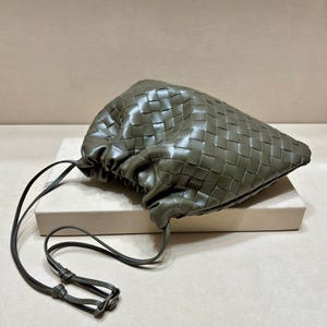May include: An olive green pouch bag featuring a woven leather exterior with a gathered drawstring closure. The bag has a long, thin matching leather shoulder strap with a small metal adjustment component at the end of the cord.