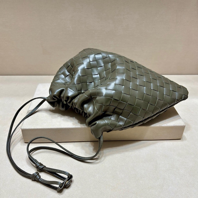 May include: An olive green pouch bag featuring a woven leather exterior with a gathered drawstring closure. The bag has a long, thin matching leather shoulder strap with a small metal adjustment component at the end of the cord.