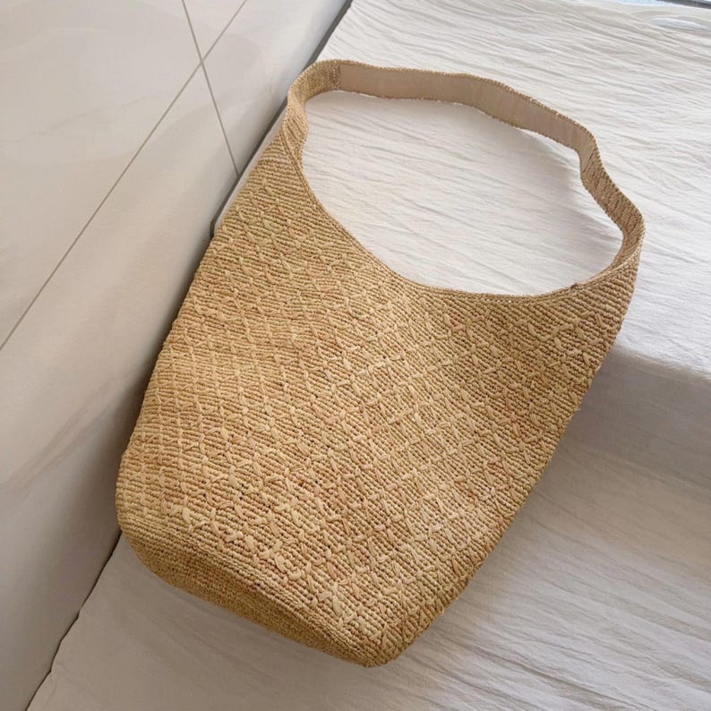 May include: A woven straw shoulder bag in a natural beige color. The bag has a wide, single strap and a textured, diamond-patterned weave. The bag is empty and sits on a white surface.