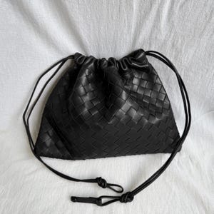 May include: A black pouch bag featuring a woven leather exterior with a grid pattern. The bag has a drawstring closure at the top that creates a gathered, ruffled edge. Two thin black leather cords extend from the sides to form a shoulder strap.