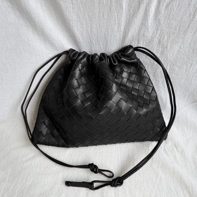 May include: A black pouch bag featuring a woven leather exterior with a grid pattern. The bag has a drawstring closure at the top that creates a gathered, ruffled edge. Two thin black leather cords extend from the sides to form a shoulder strap.