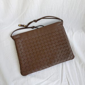 May include: A brown leather shoulder bag featuring a woven basket-weave pattern across the front panel. The bag has a smooth leather top border, a thin adjustable shoulder strap with gold-toned metal hardware, and a rectangular silhouette.