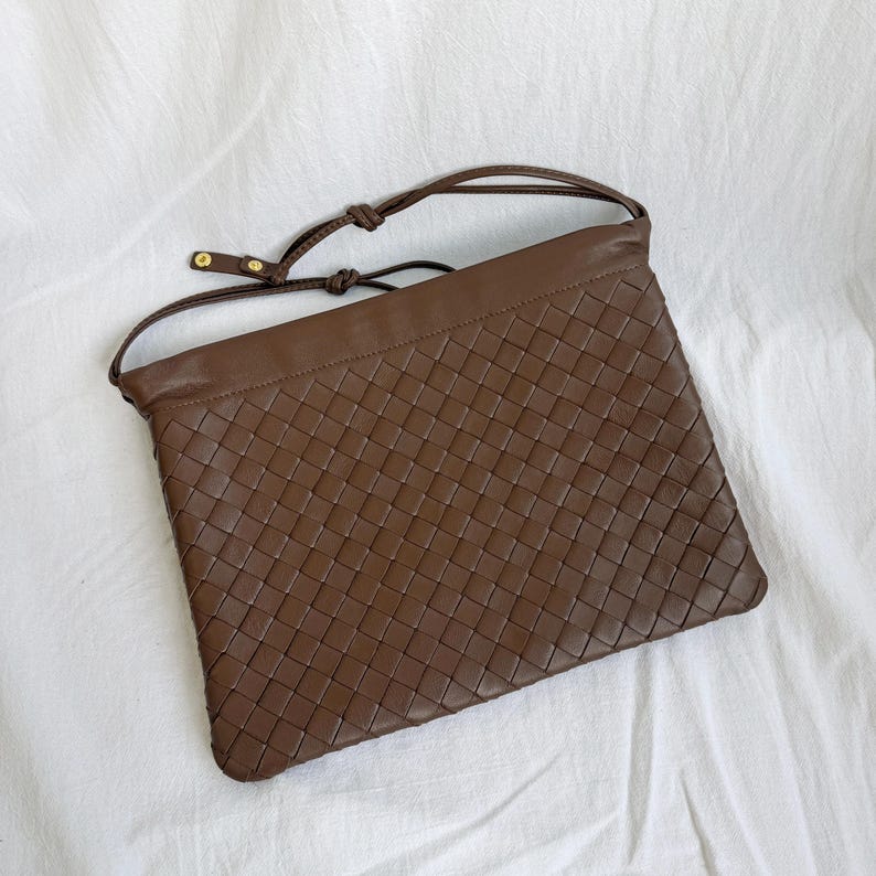 May include: A brown leather shoulder bag featuring a woven basket-weave pattern across the front panel. The bag has a smooth leather top border, a thin adjustable shoulder strap with gold-toned metal hardware, and a rectangular silhouette.