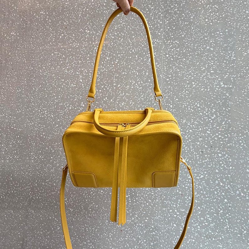 K&ouml;nnte beinhalten: Eine leuchtend gelbe Wildlederhandtasche mit rechteckiger Silhouette, einem Rei&szlig;verschluss oben mit langen Lederzuglaschen, zwei kurzen Tragegriffen und einem langen, verstellbaren Schulterriemen, der mit goldfarbenen Metallbeschl&auml;gen befestigt ist.
