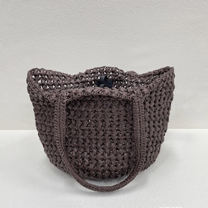 May include: A woven tote bag in a dark brown hue. The bag features a textured, open weave design with a rounded handle and a black clasp closure. The bag's shape is a structured, open-top design.