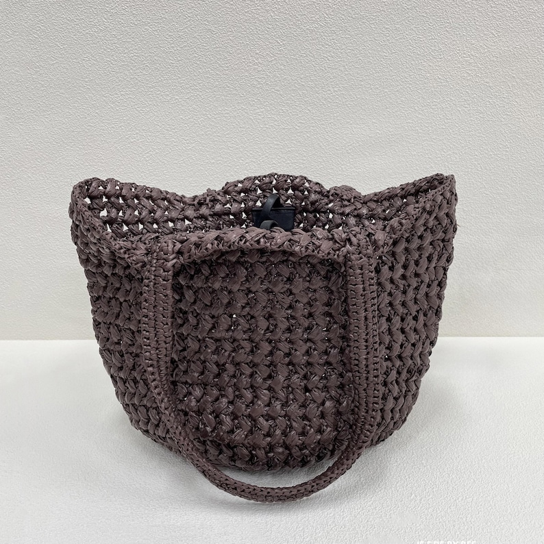 May include: A woven tote bag in a dark brown hue. The bag features a textured, open weave design with a rounded handle and a black clasp closure. The bag's shape is a structured, open-top design.