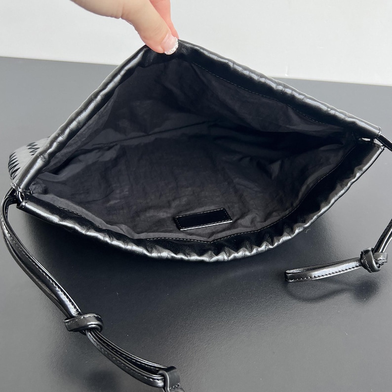 May include: A black shoulder bag with a smooth, slightly gathered exterior and a matching black interior lining. The bag features a thin, adjustable black strap with knotted ends and a small, rectangular black label sewn inside the main compartment.