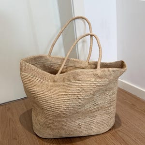May include: A large, open-top tote bag made of woven natural fibers. The bag has two sturdy handles and a spacious interior. The neutral color and textured weave give it a casual, beachy aesthetic. The bag is designed for carrying items.